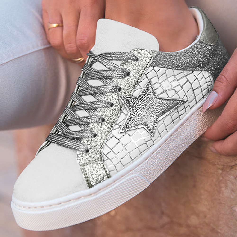 A close-up of a person wearing a white sneaker with metallic silver textured panels, a silver star detail, and matching silver laces, while sitting in white pants.